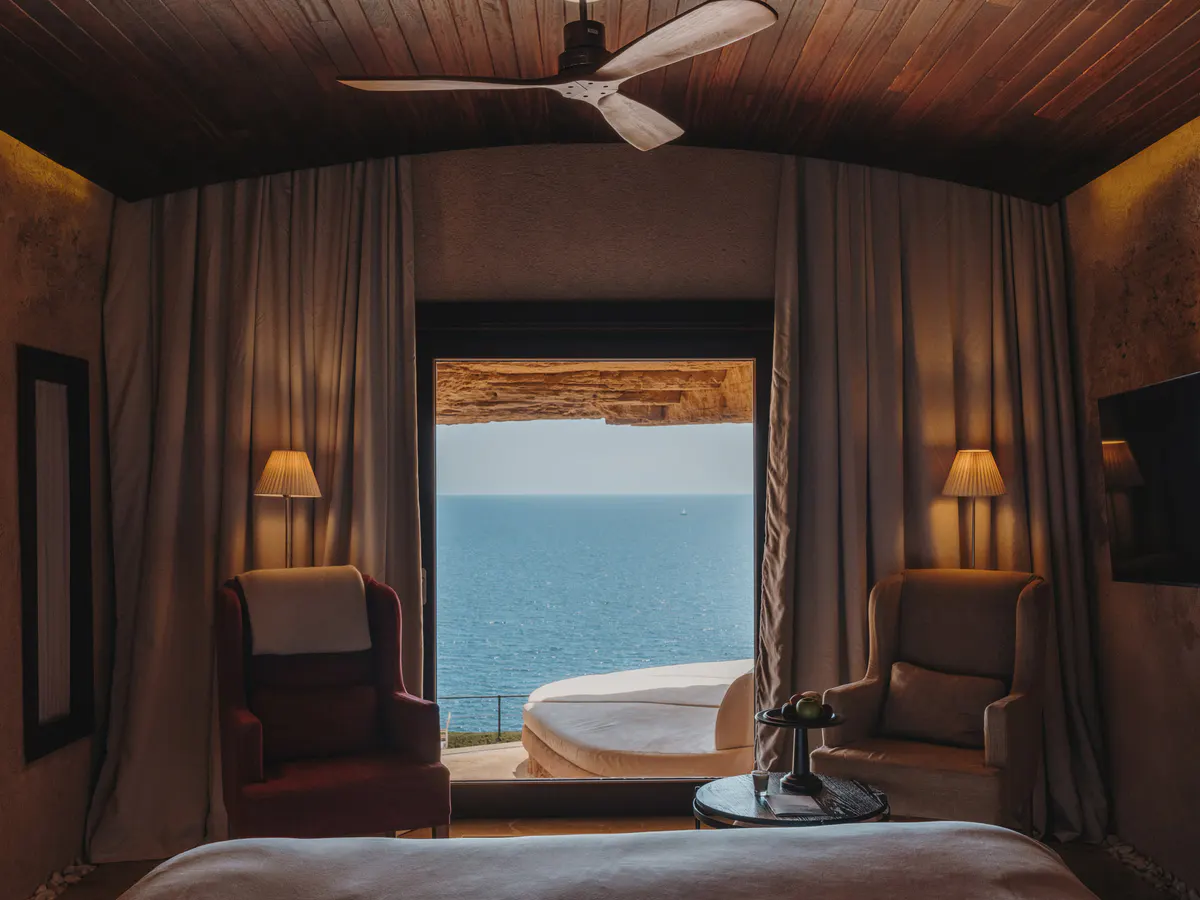 A curtained bedroom framing turquoise water through an arched window, with gold sconces and lounge chairs