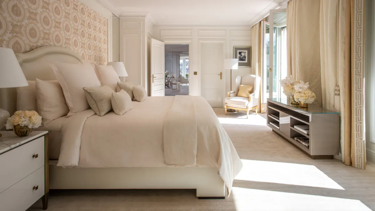 A bedroom with textured headboard, cream linens, and soft light from tall windows