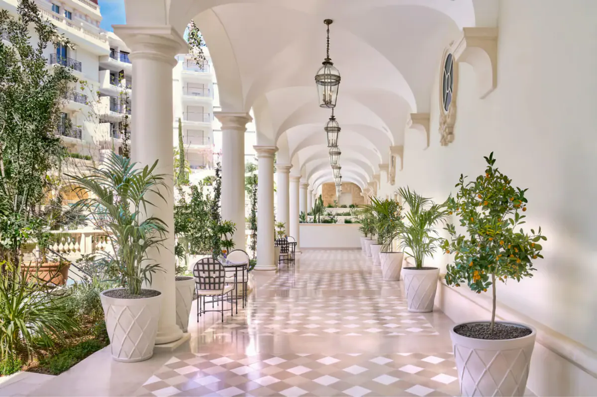 Covered colonnade walkway with white columns, potted plants lining both sides, and sunlight casting geometric shadows