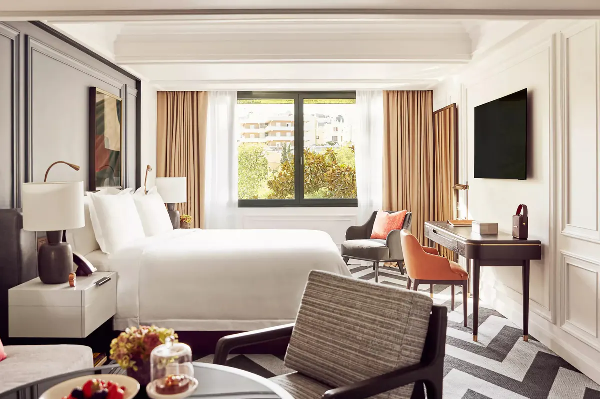 A bedroom with white linens, window seating, and warm gold curtains with city views