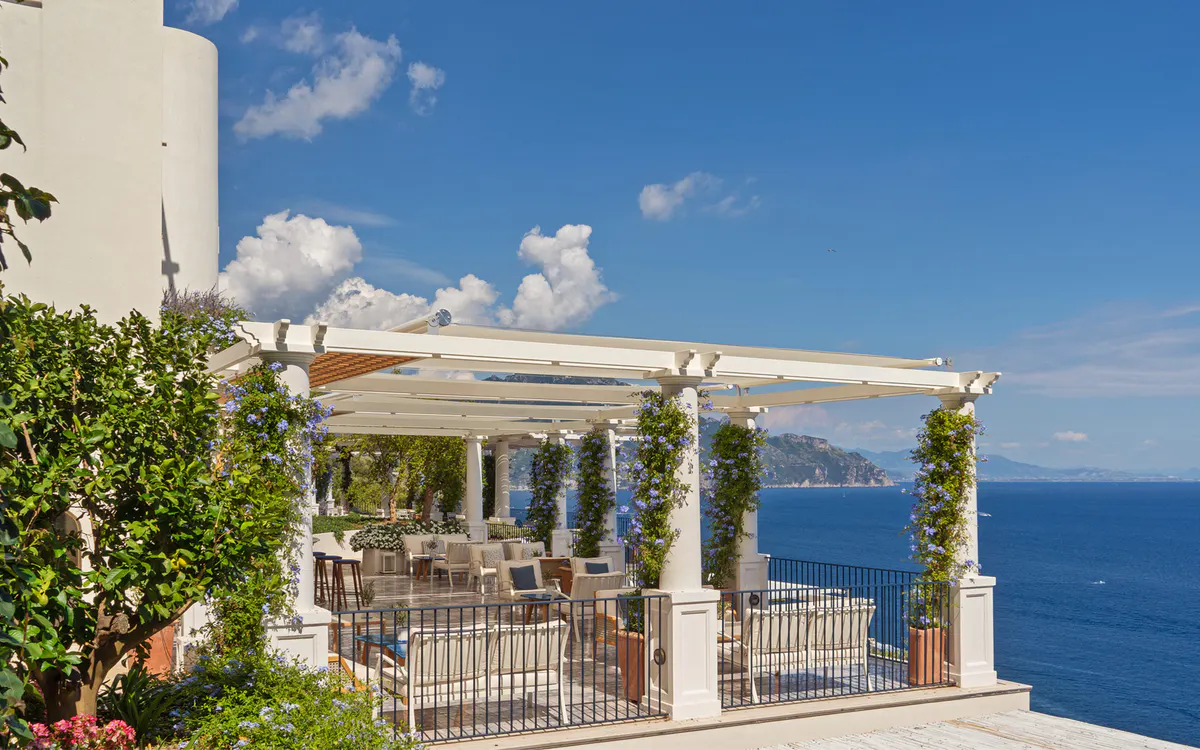 Pergola dining area with white posts supporting climbing vines overlooking blue sea