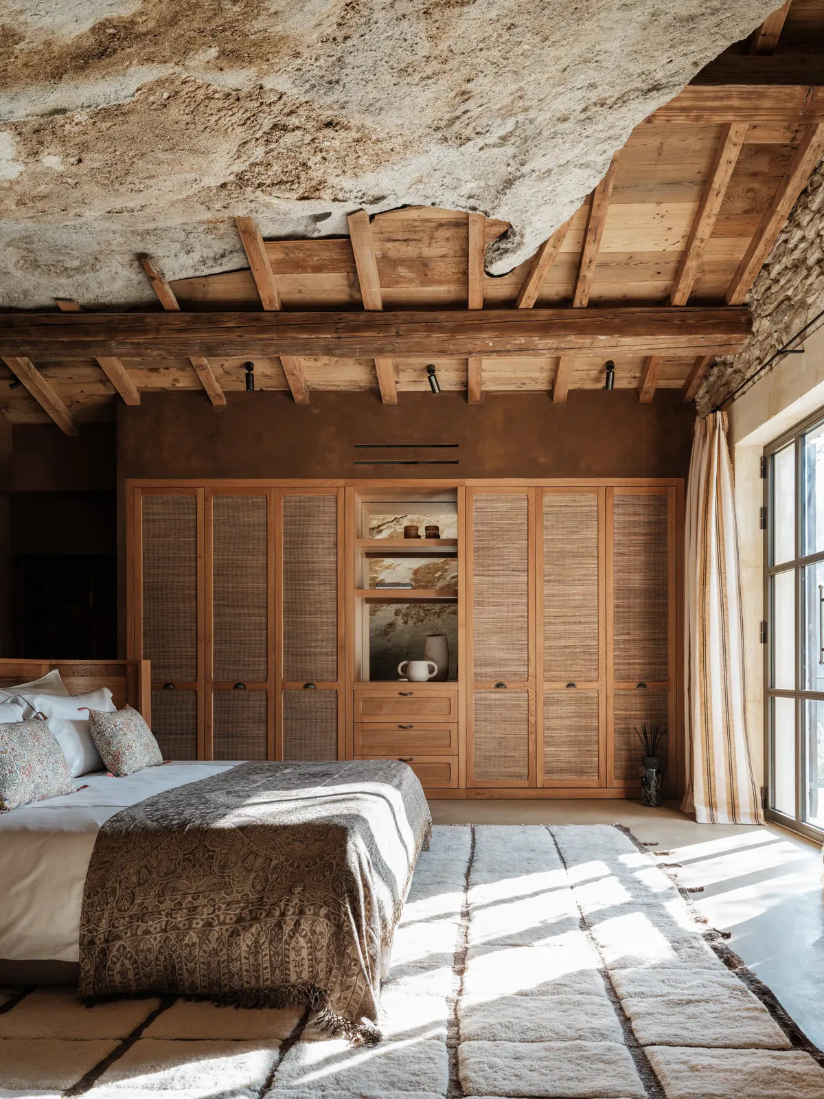 Rock-hewn bedroom with dramatic raw limestone ceiling, wooden structural beams, timber wardrobe cabinet