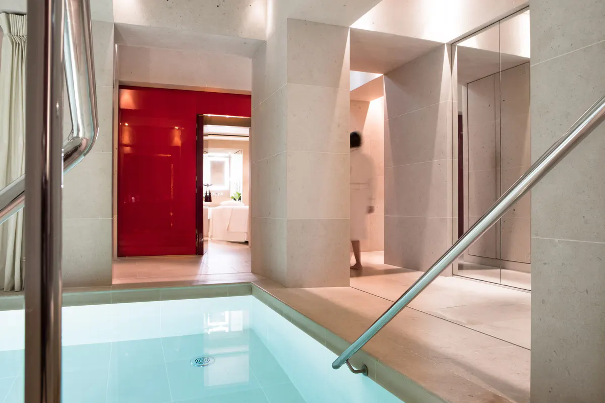 A spa corridor with glass partitions, red walls, and a soaking tub visible through doorway