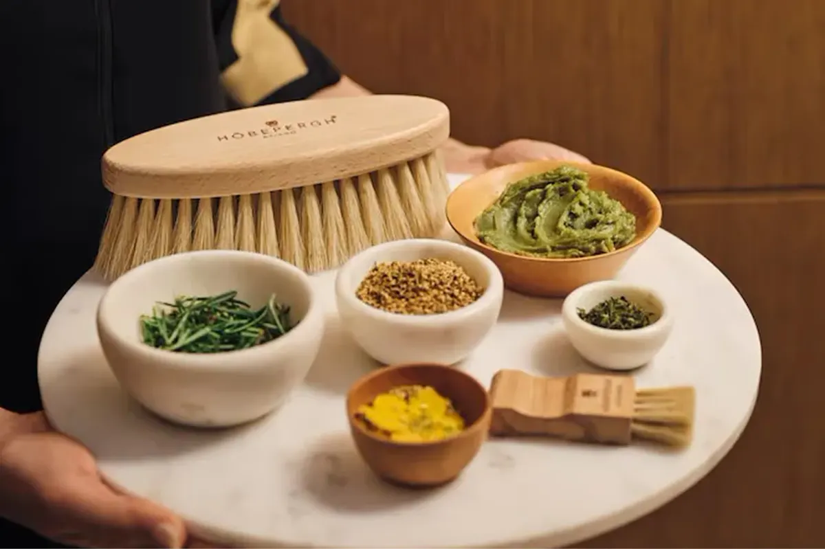 Spa treatment items in white bowls on a table with natural ingredients and a wooden brush