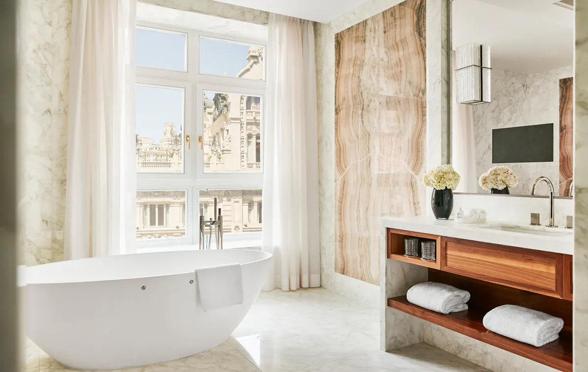 Spa bathroom with freestanding tub, marble surround, wood vanity, and arched window with city views