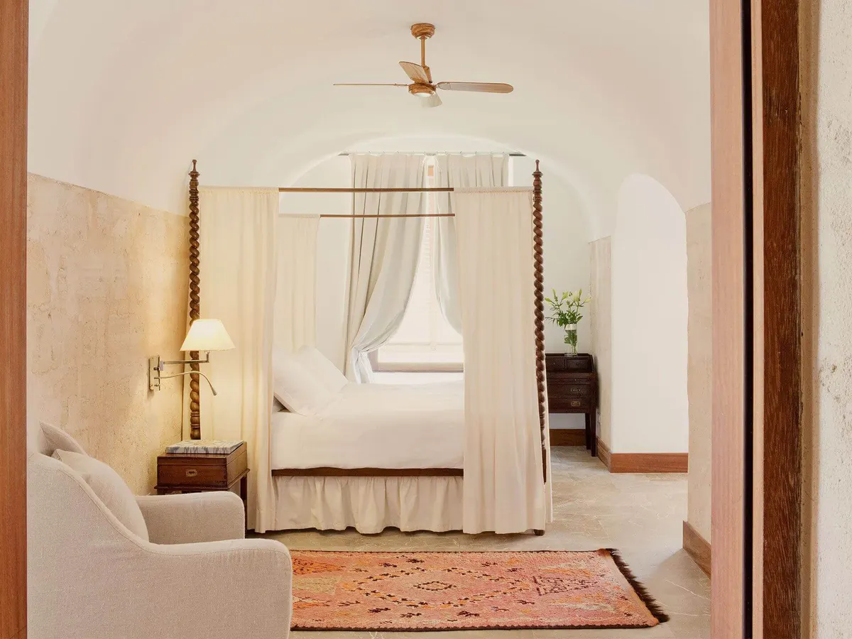 A window draped in cream linen overlooks pale grass and turquoise sea at dusk