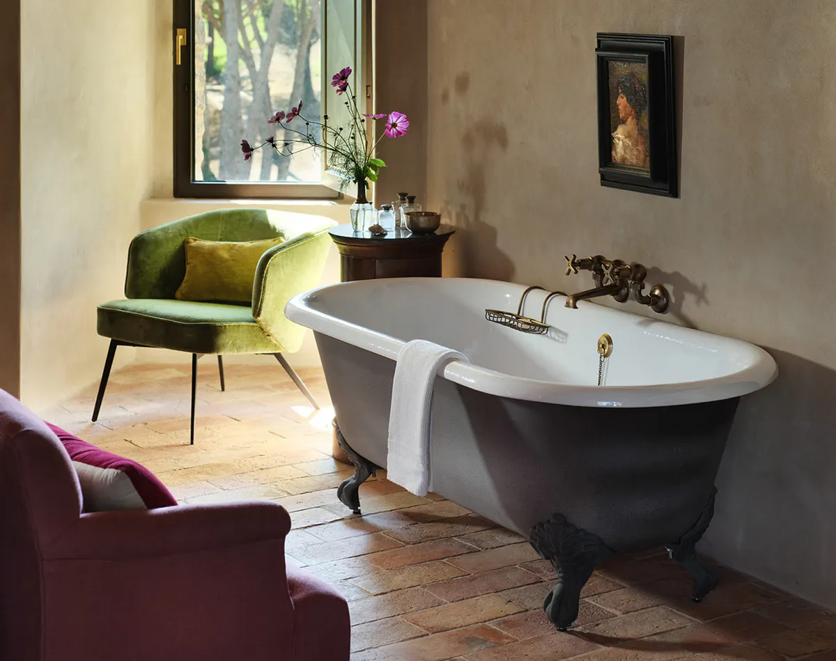 A bathroom with a white clawfoot tub beside an arched window, an olive velvet armchair, and terracotta tile