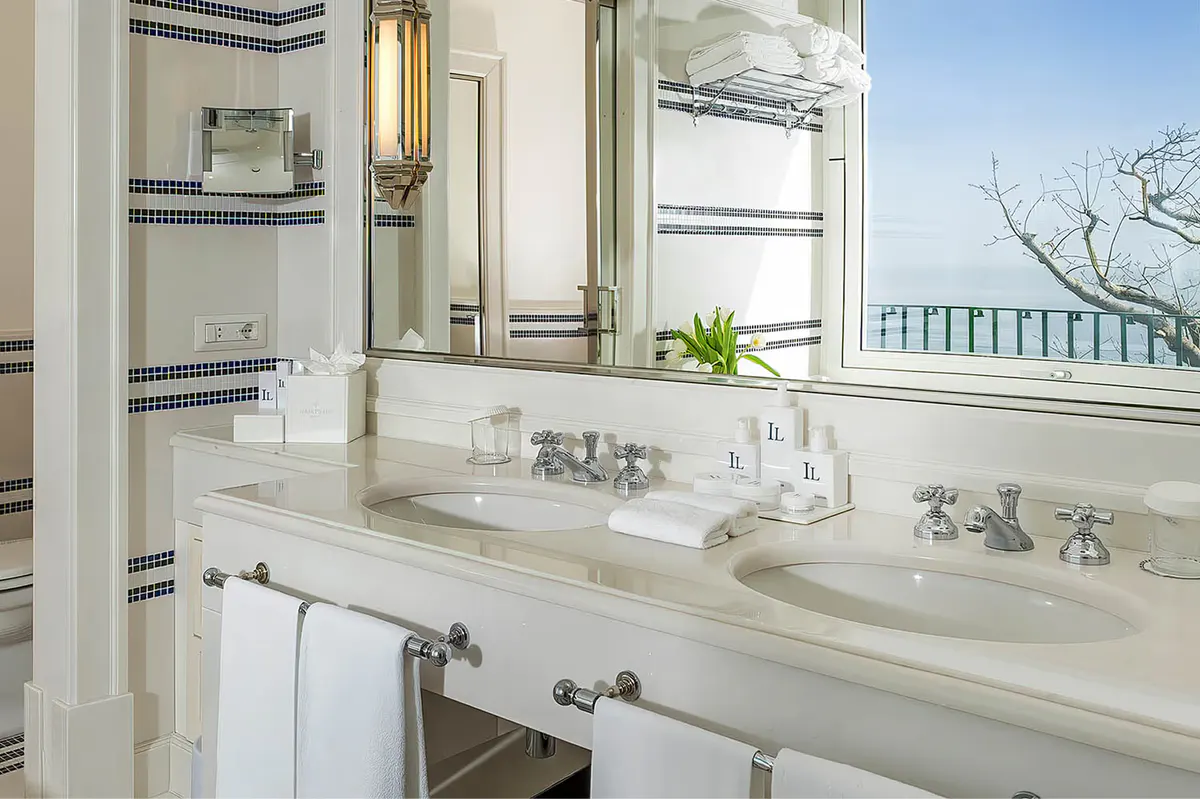 Marble vanity with dual sinks, brass fixtures, windows overlooking Mediterranean, white tile surround