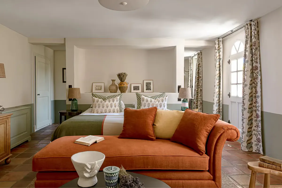 A spacious room with pale green walls, a daybed lounge, patterned curtains, and bright windows overlooking countryside