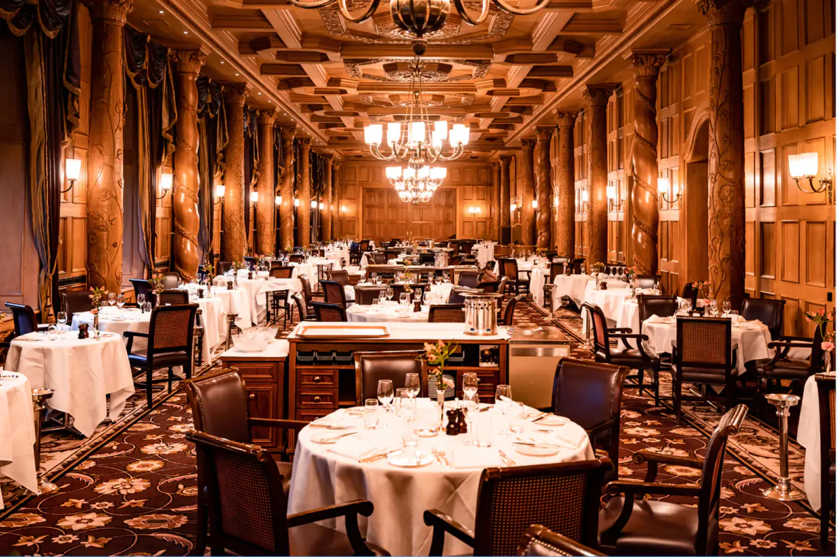 A grand dining hall with ornate golden ceiling, crystal chandeliers, and formally set white-draped tables