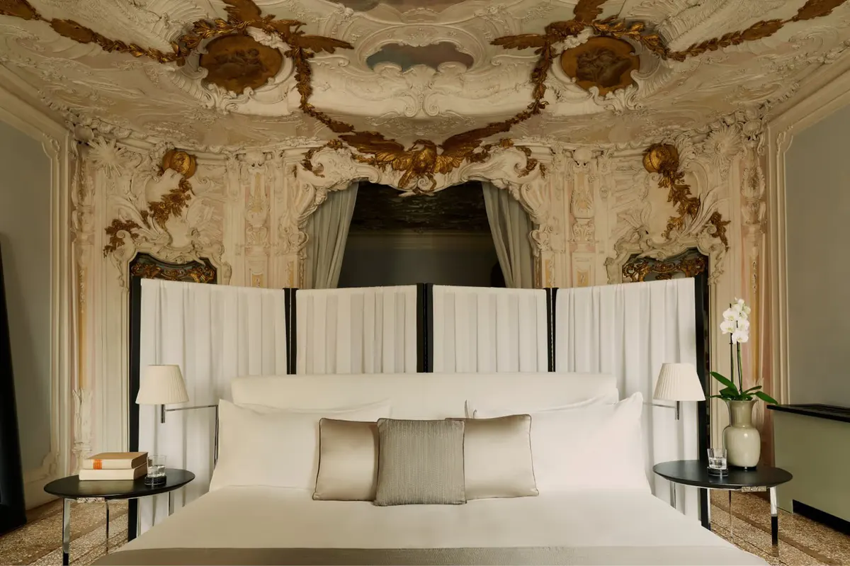 A bedroom with ornate frescoed ceiling, four-poster bed with white bedding, and matching carved wooden nightstands