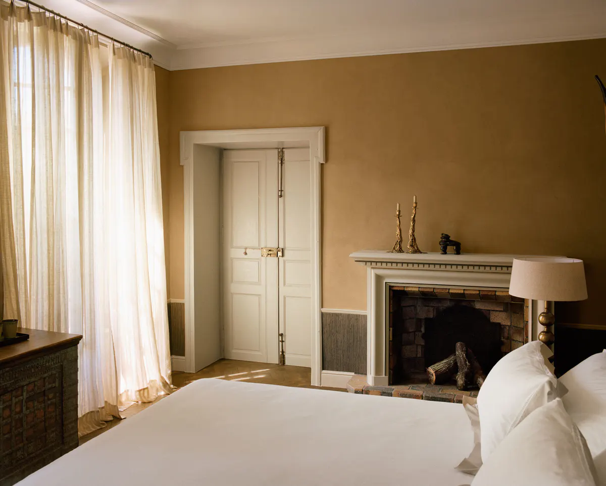 Bedroom with fireplace mantel, multiple windows with flowing curtains, and warm earth tones