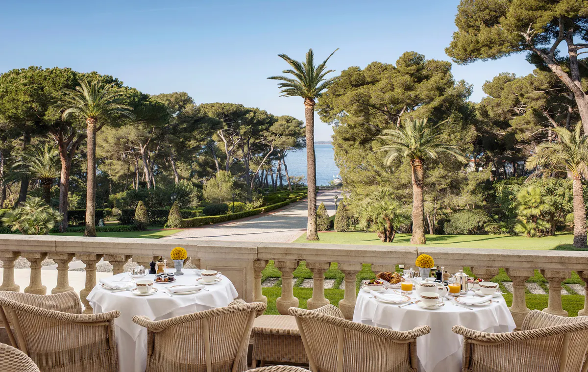An outdoor dining table set on a terrace with palm trees and sea views beyond manicured lawns