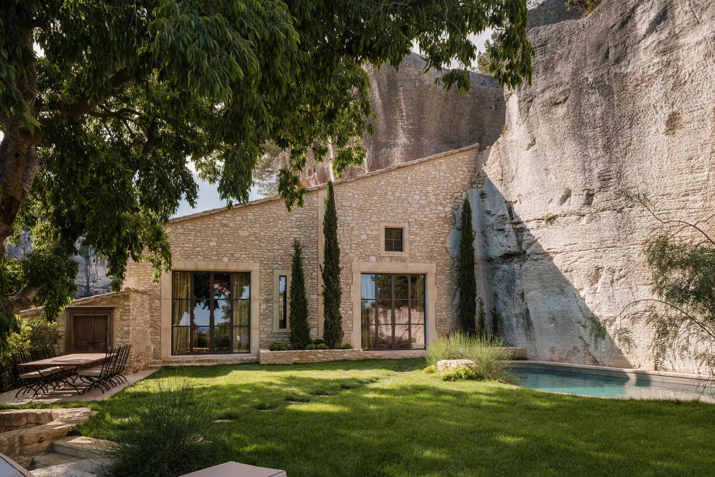 Stone villa with modern glass doors, wooden pergola, grass courtyard, pale turquoise pool, limestone cliff backdrop