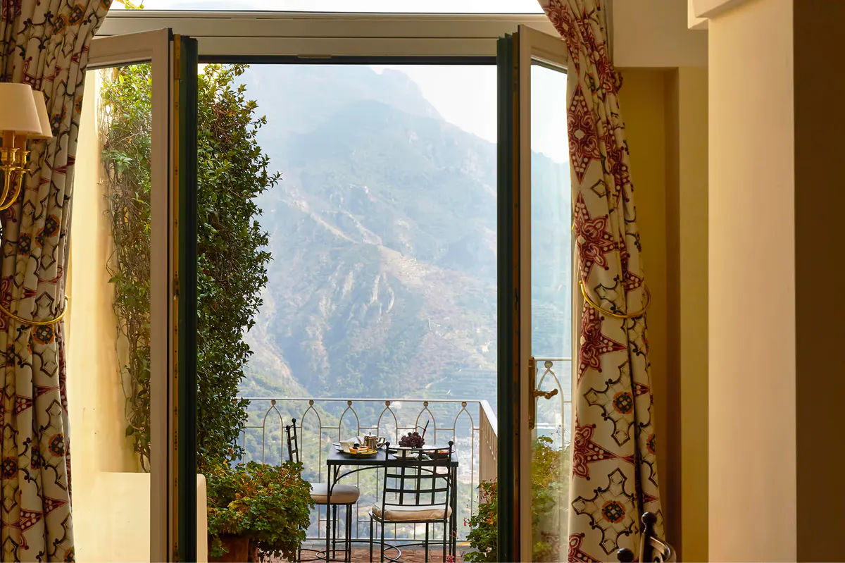 Room window framing coastal mountains with floral patterned curtains and wrought-iron balcony railing